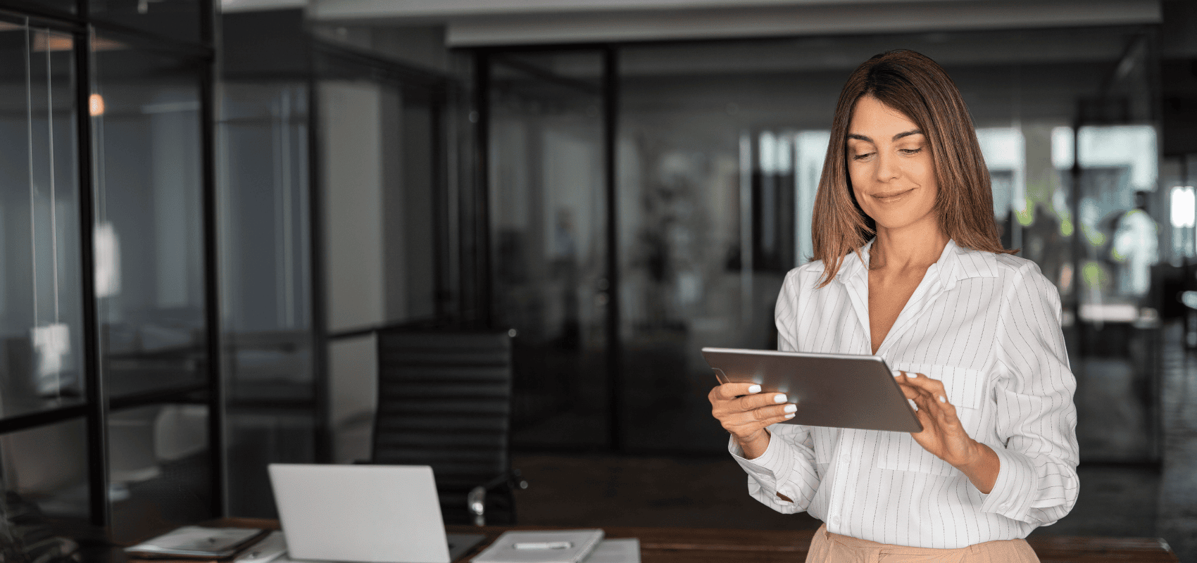 A woman in a white striped shirt using a tablet in a modern office with a laptop and papers on the table.