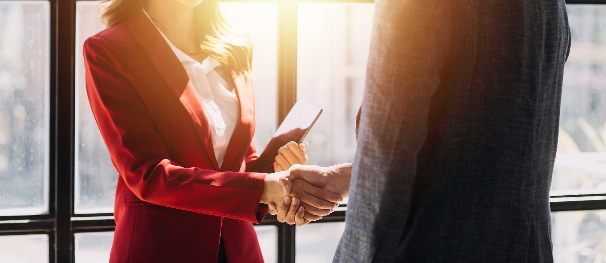 A woman in a red blazer shakes hands with a man in a suit, sunlight streaming through a window.