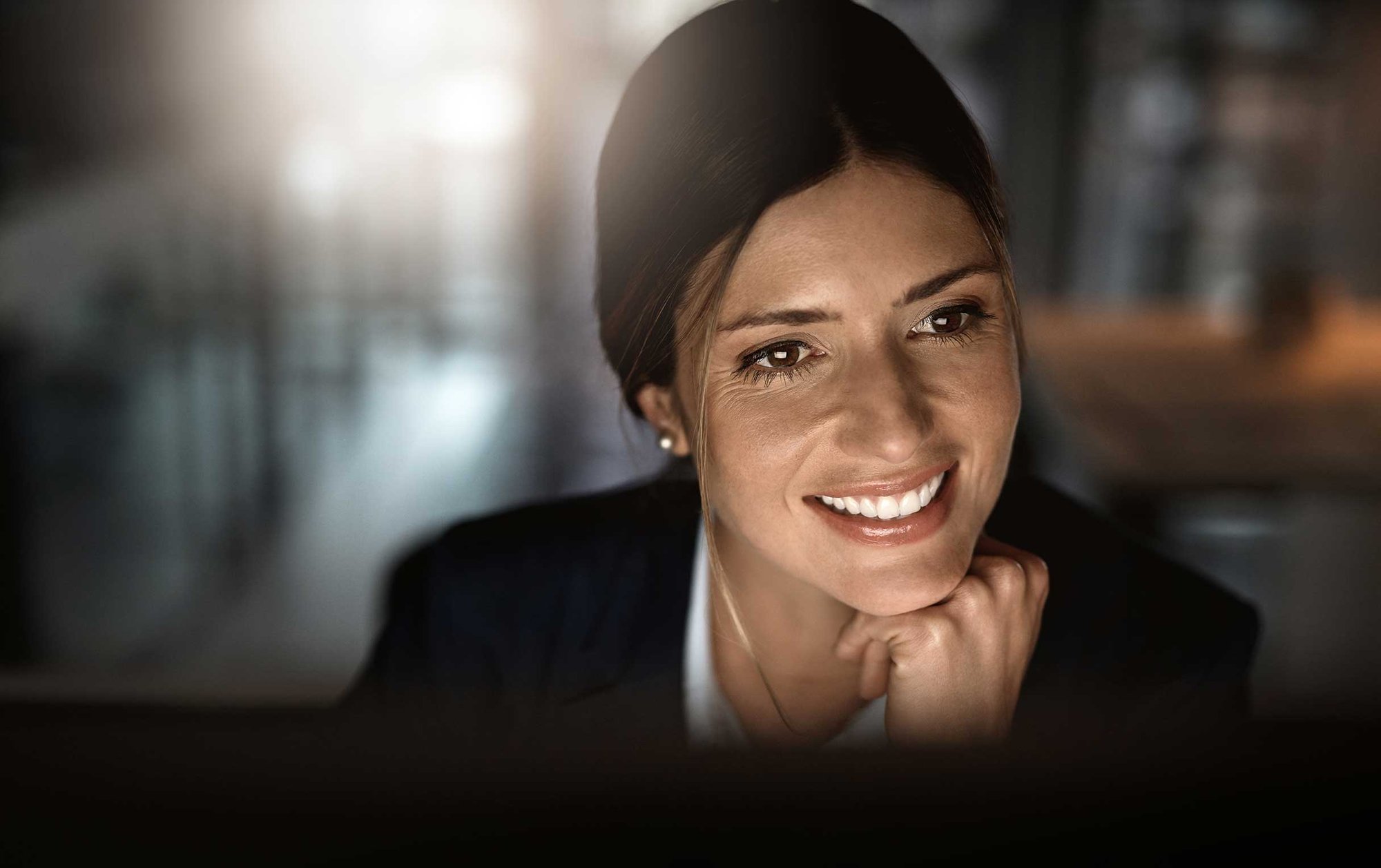 A smiling woman with long brown hair rests her chin on her hand, looking warmly at a screen.