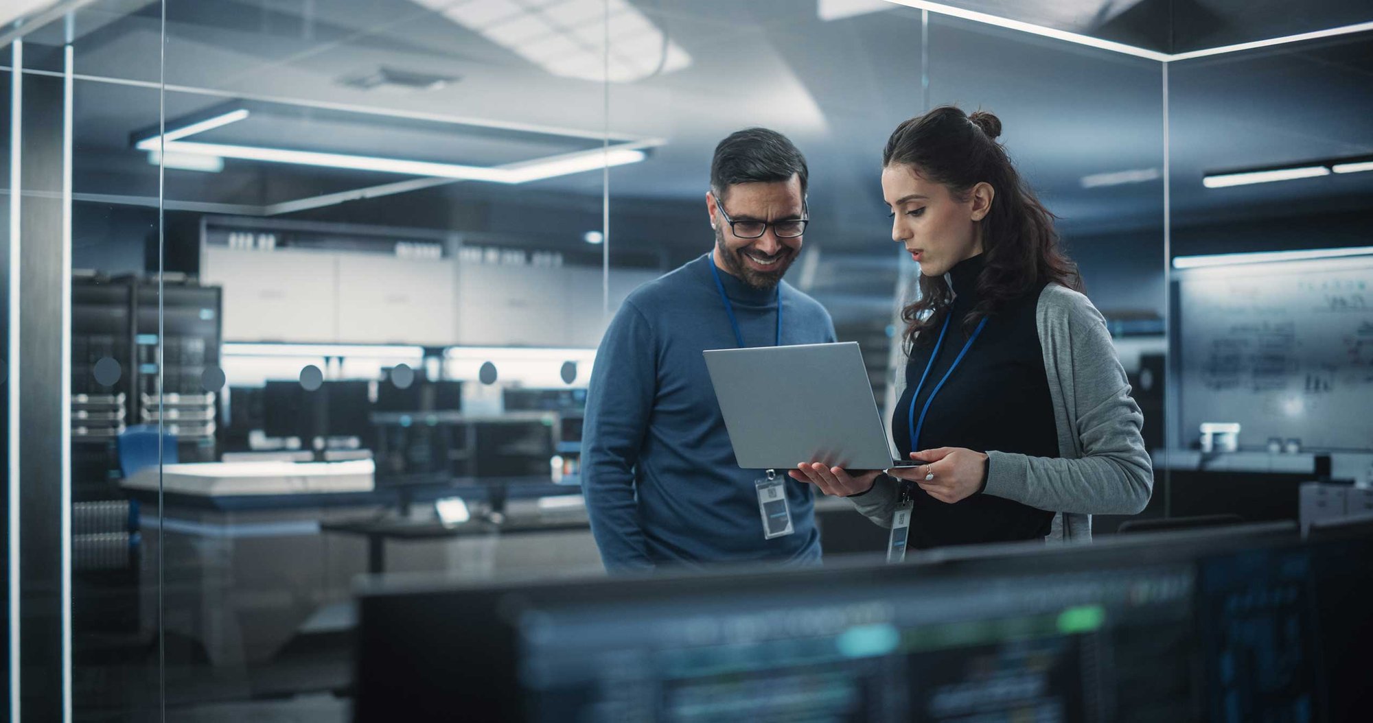Two professionals in an office discussing information on a laptop, with computers in the background.