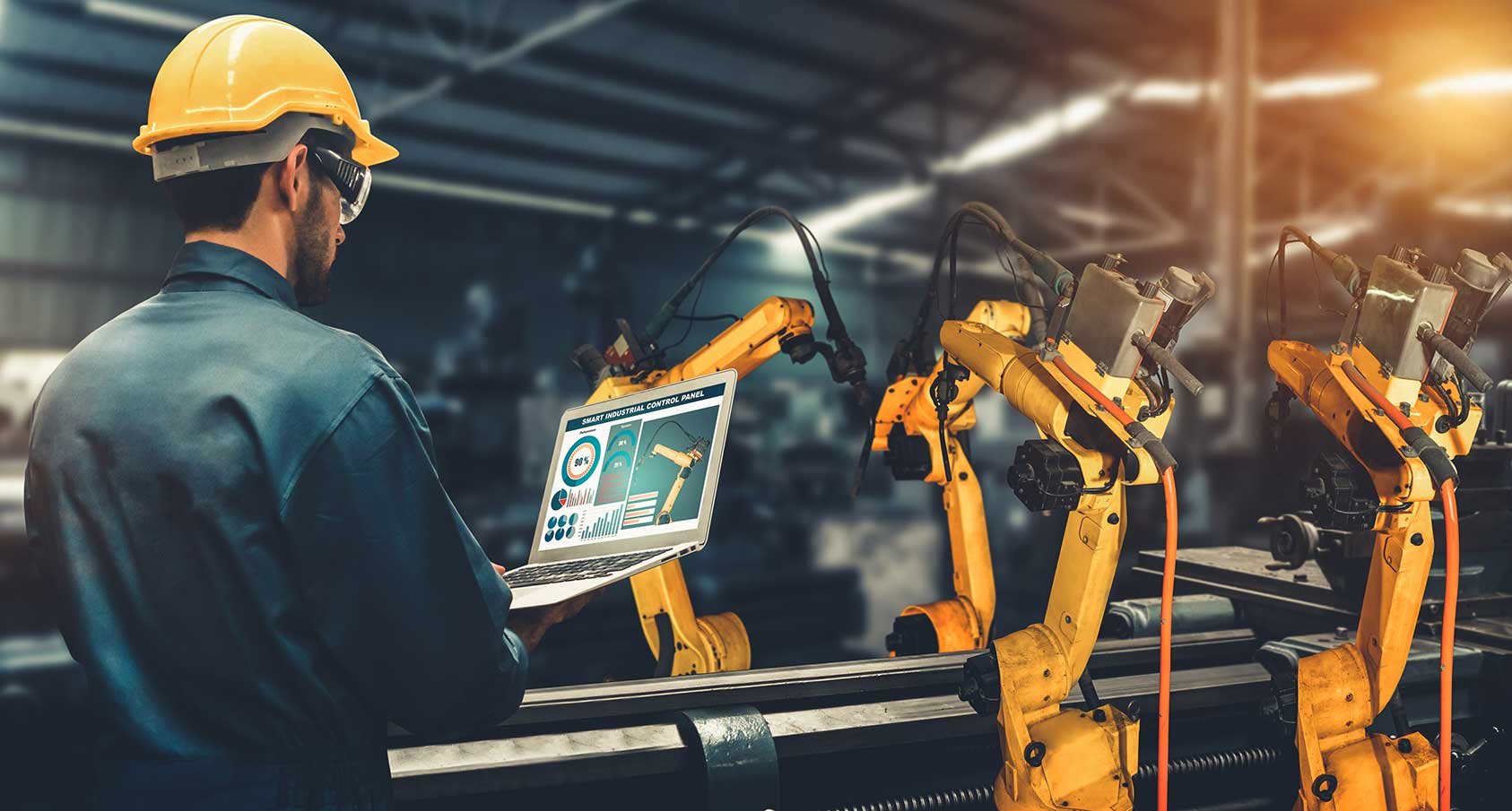 A worker in a hard hat uses a laptop to control yellow robotic arms in a factory.
