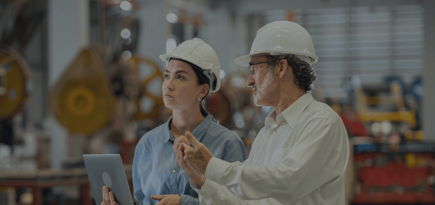A woman in a hard hat holds a laptop, listening to a man in glasses and a hard hat talking in a workshop.